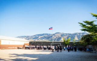 School yard with children playing soccer, Sunny clear blue sky, mountain landscape, and Haitian flag in background.