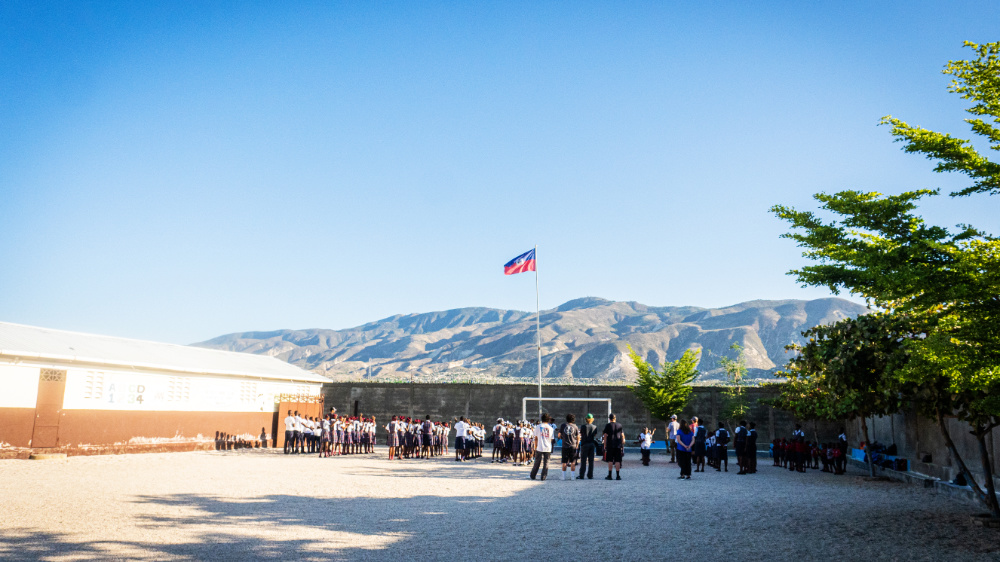 School yard with children playing soccer, Sunny clear blue sky, mountain landscape, and Haitian flag in background.