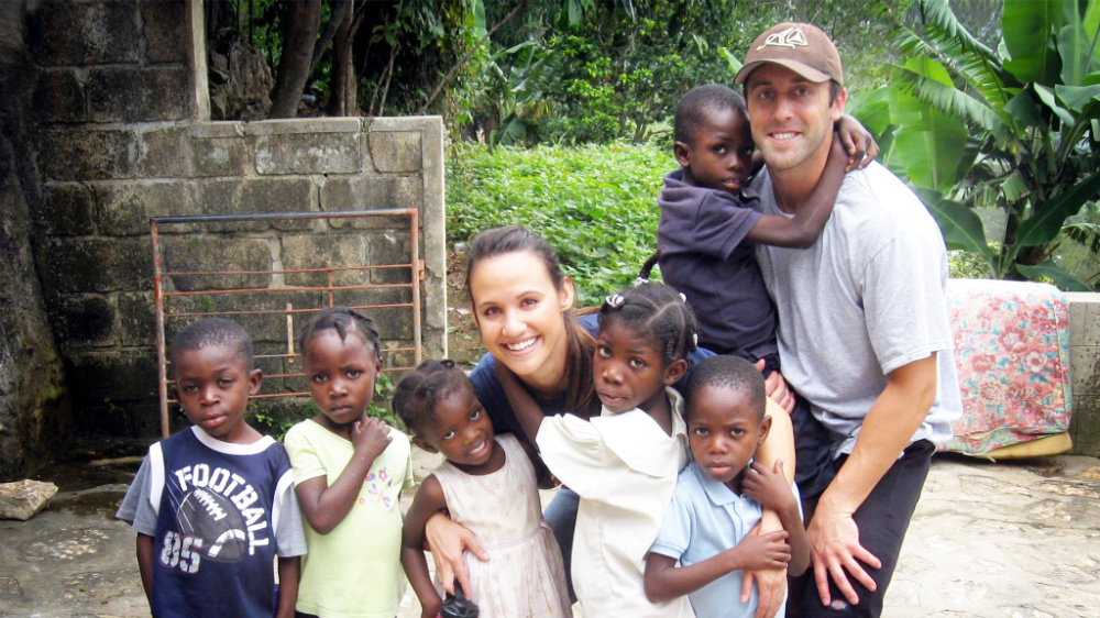 A woman and man volunteer with small Haitian children smiling and posing for a picture.