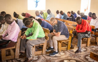 Men sitting on wood benches with heads bowed in church praying.