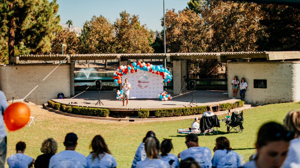 Simi walk speaker on a grand stage decorated in balloons in the park with a fountain and lake in the background.