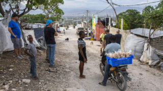 People by a dirt road buying good from a merchant on a motorcycle.