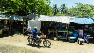 Vendors along a dirt road, motorcyclist using phone nearby.